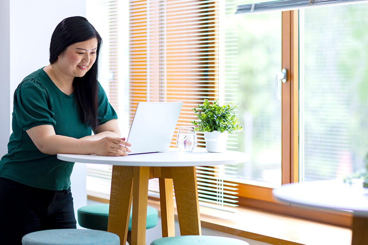 A student in a green shirt uses a laptop on a round table by a sunny window. A small potted plant and glass of water are beside her.